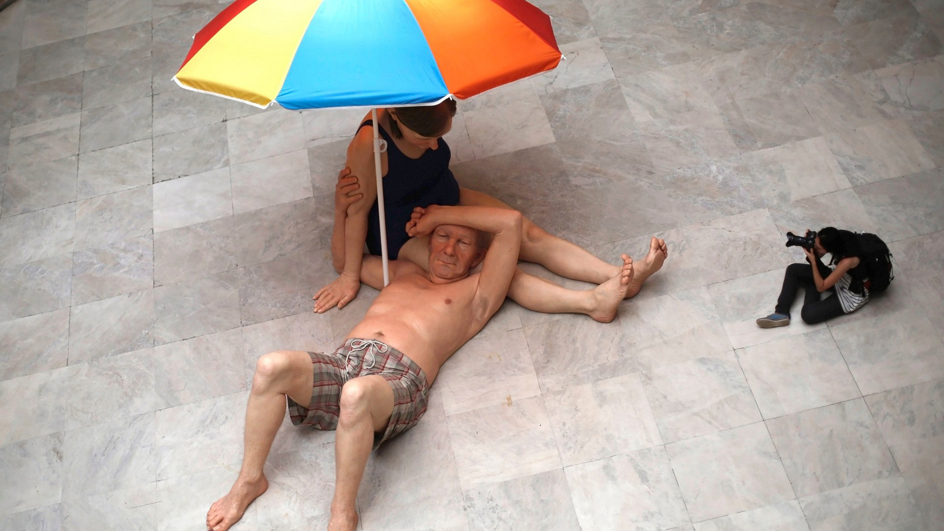 Sculpture Couple Under an Umbrella by Ron Mueck showing two figures huddled together under shelter, expressing intimacy, care, and protection.